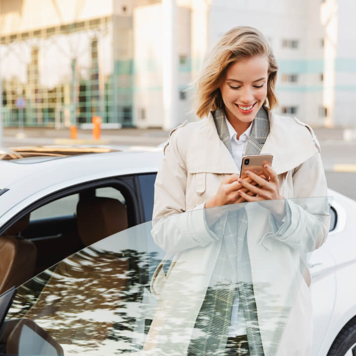 A smiling woman checking her phone while standing next to her car on a street in Toronto.