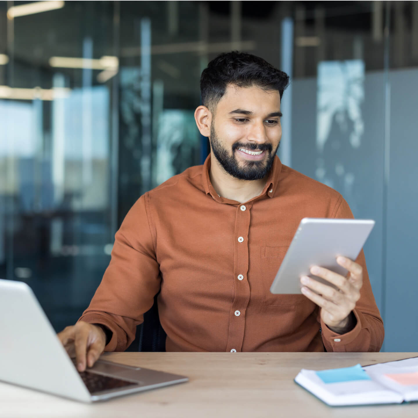 A smiling man in an orange shirt is looking at a tablet in his hand and using a laptop computer.