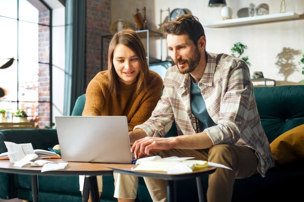 S'occuper de son assurance chez soi : un couple heureux utilise un ordinateur portable, assis sur un sofa dans un appartement
