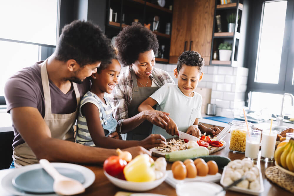 A happy family of four prepares healthy food in the kitchen. The mother helps the son cut tomatoes while the daughter watches in the arms of the father.