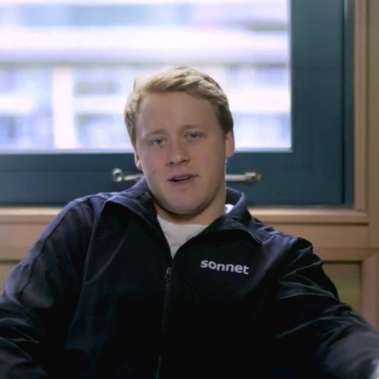 Morgan Rielly of the Toronto Maple Leafs seated indoors wearing a black jacket with Sonnet logo, positioned in front of a window with a blurred cityscape in the background.