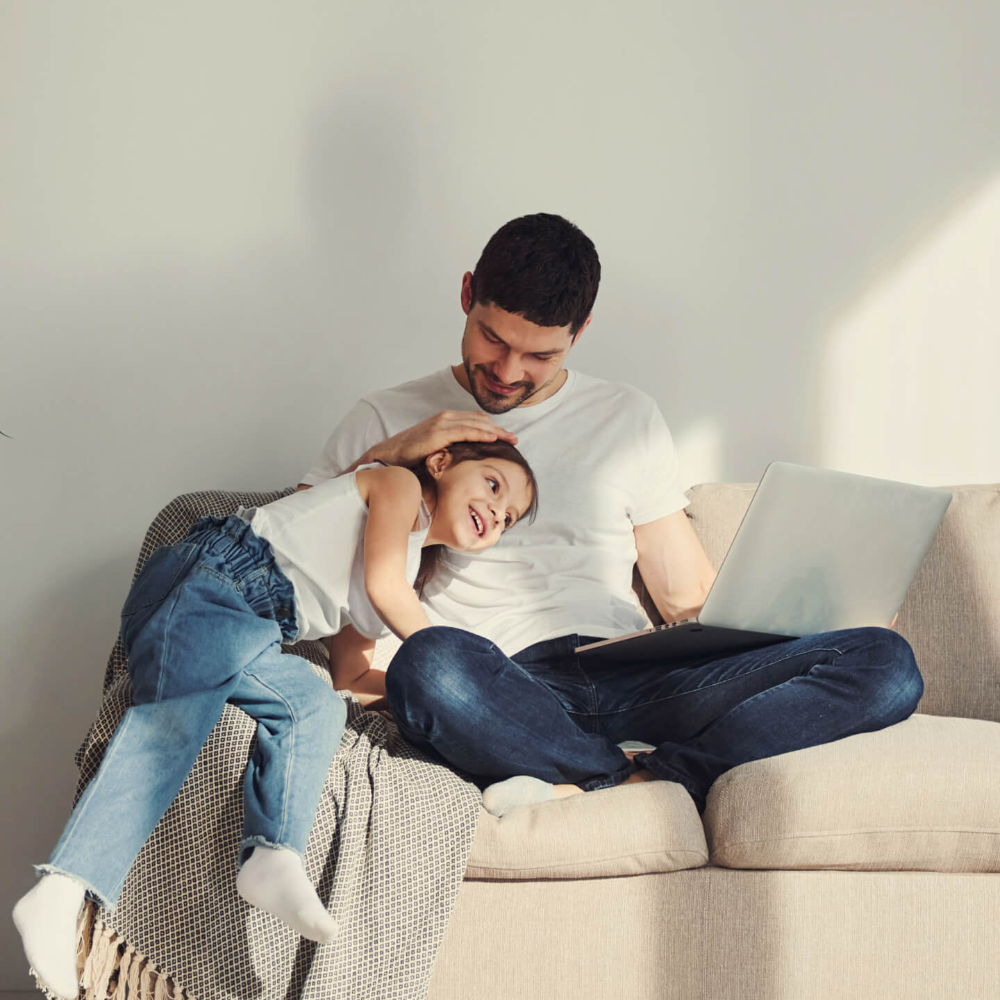 A father sits on a couch and his daughter sits on the arm rest with her head on his chest while he lovingly embraces her.