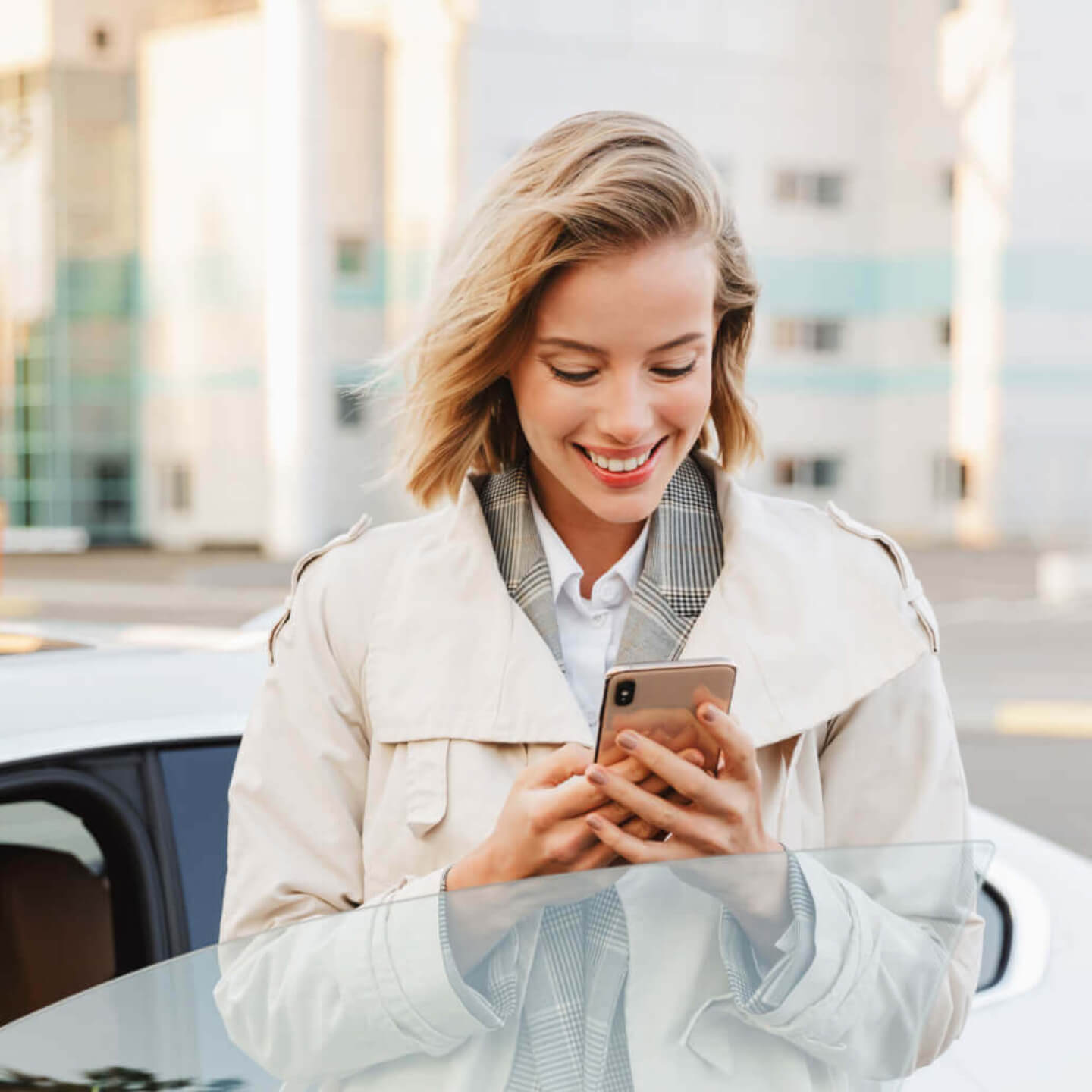 Une femme sourit en regardant son téléphone cellulaire, debout, près de sa voiture, sur une rue de Canada.