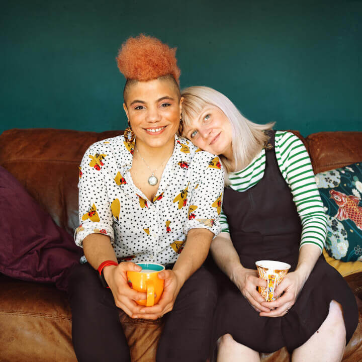 Two smiling women sitting together on a couch, one woman leans her head on the other’s shoulder.