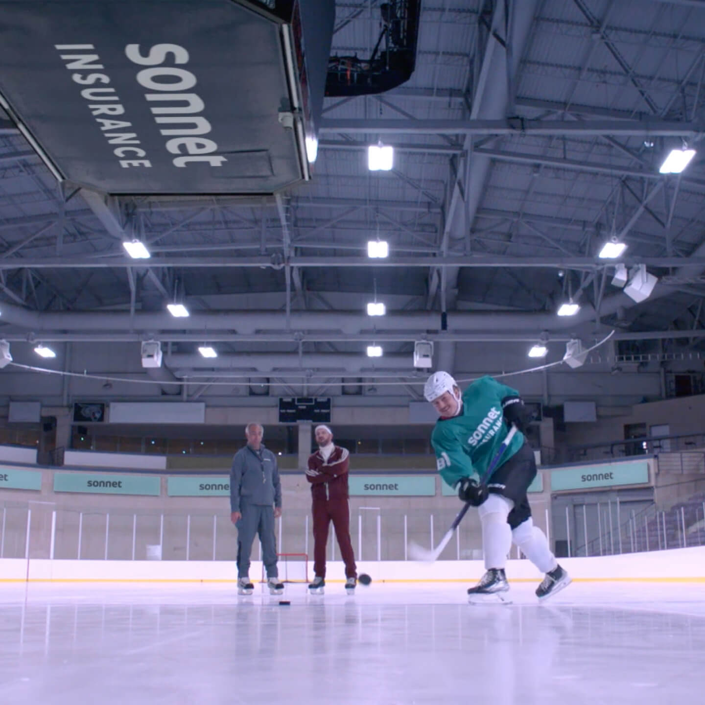 William Nylander of the Toronto Maple Leafs skating on an indoor ice rink wearing a teal Sonnet Insurance jersey, with two coaches standing nearby and a Sonnet-branded scoreboard overhead.