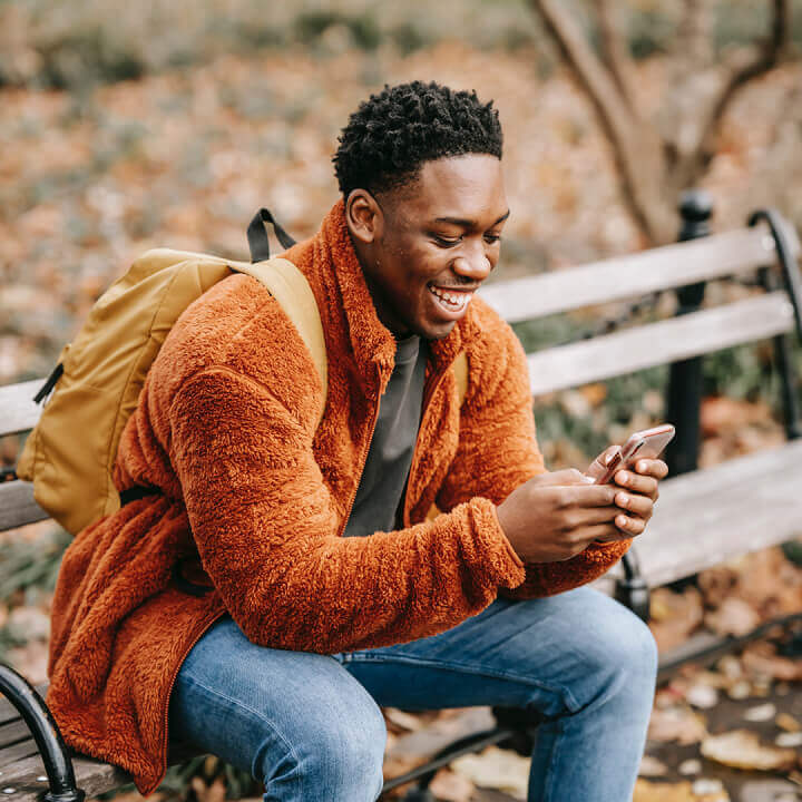 Un homme souriant avec un sac à dos assis à l’extérieur sur un banc de parc regardant son téléphone.