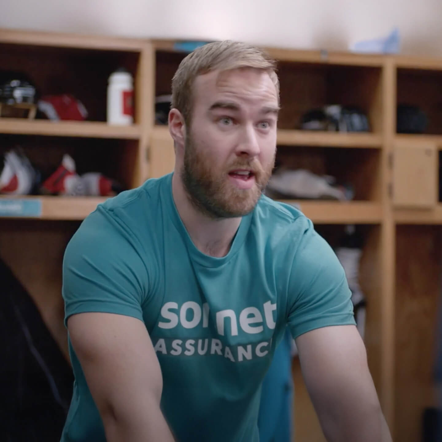 David Savard des Canadiens de Montréal porte un T-shirt Sonnet Assurance sarcelle, il est assis dans un vestiaire avec des cases en bois et des équipements sportifs en arrière-plan.