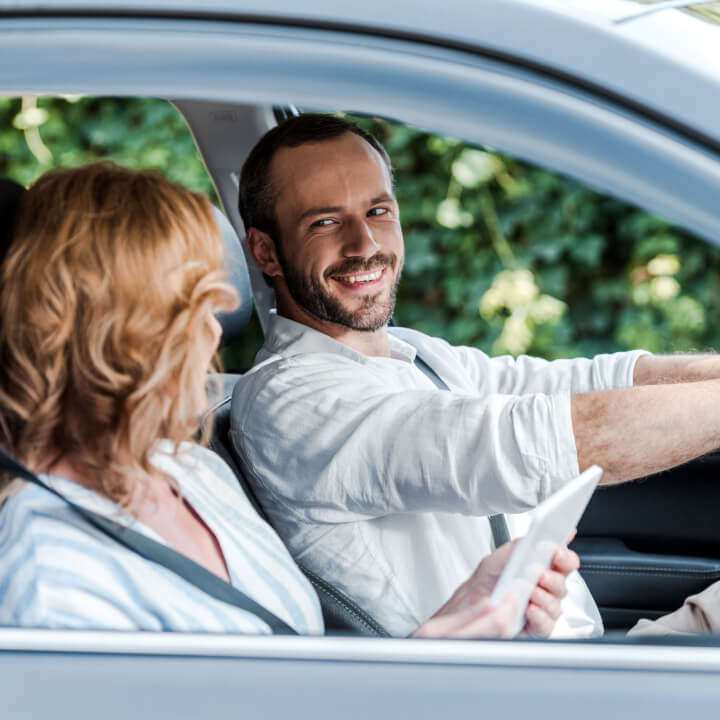 Happy man driving a car, looking at his wife in the passenger seat who is using a tablet to find directions around Canada.