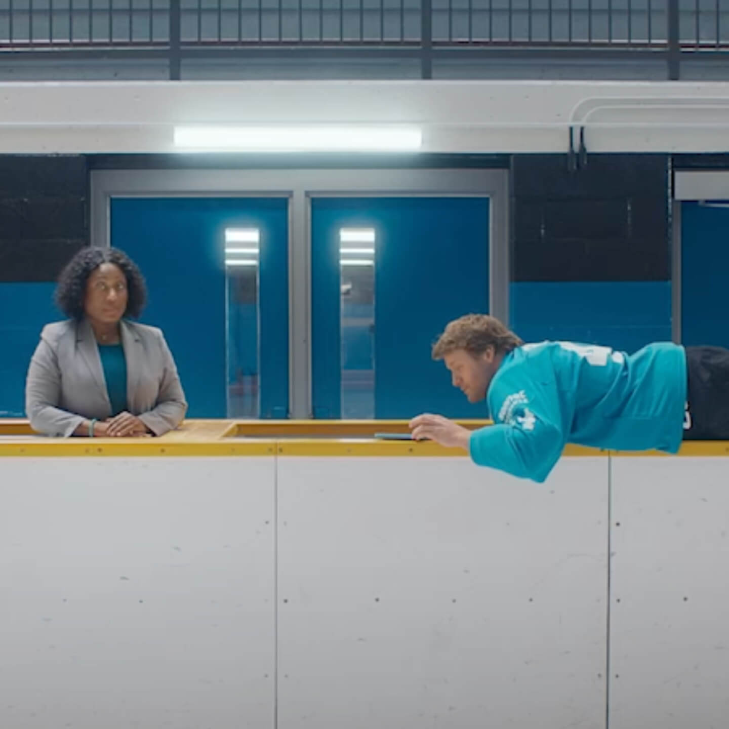 Morgan Rielly of the Toronto Maple Leafs wears a teal hockey jersey as he uses his smartphone while lying across the boards of an ice rink. A woman in a gray blazer stands next to him behind the boards.