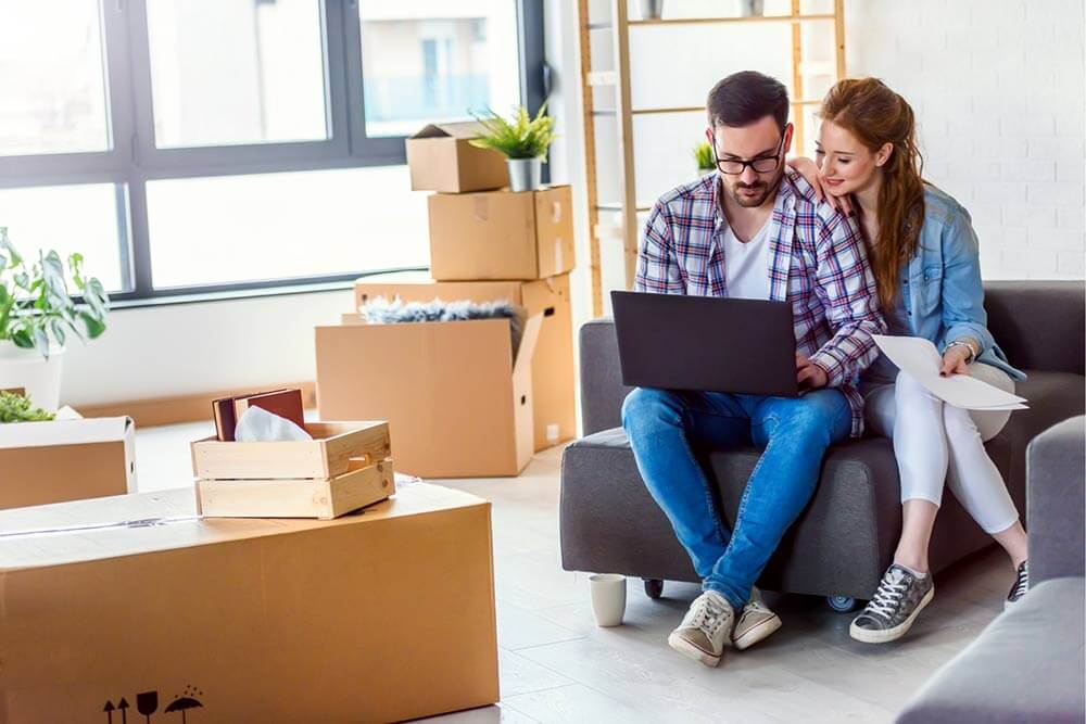 Young couple moving in a new home. Sitting and relaxing after unpacking. Searching home insurance on a laptop.