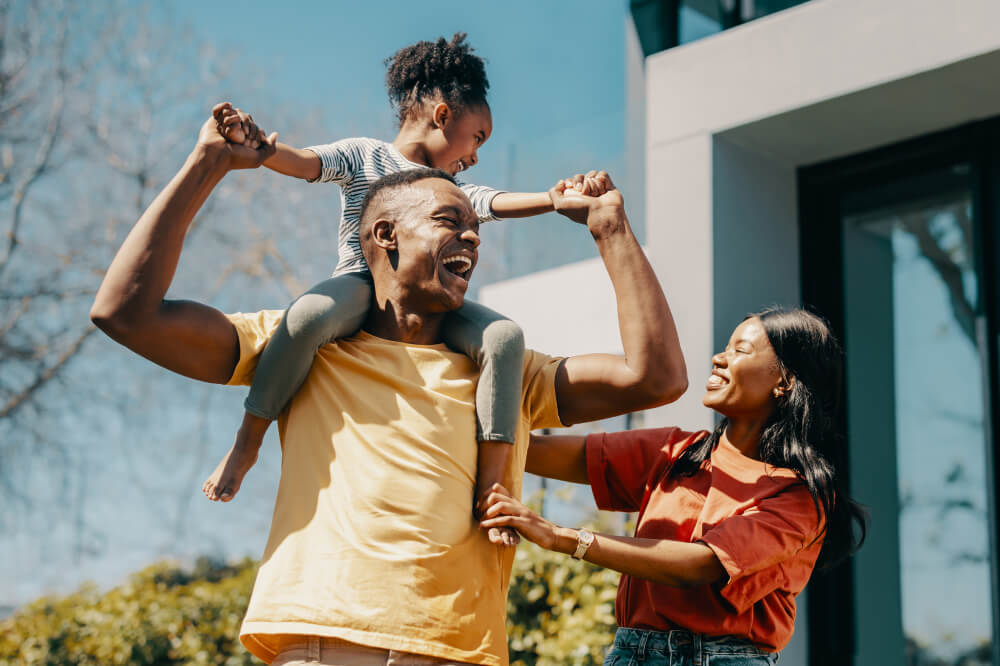 Une famille heureuse de trois personnes profite du soleil près de leur maison. La jeune fille est assise sur les épaules de son père, et la mère les regarde. Tous rient et sourient de bon cœur.