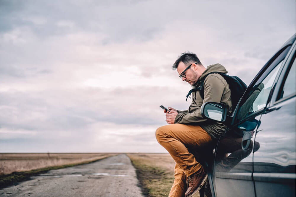 Man on a cellphone sitting on his car on an open road