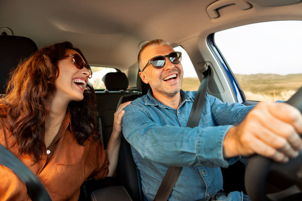 Un couple heureux se balade en voiture, lunettes de soleil aux yeux. L’homme sourit en regardant la route tandis que la femme lui sourit et pose une main sur son épaule.